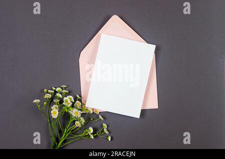 Enveloppe rose avec carte blanche vide pour le texte et le bouquet de pâquerettes sur fond gris. Maquette d'invitation ou de carte de vœux vierge avec espace de copie Banque D'Images