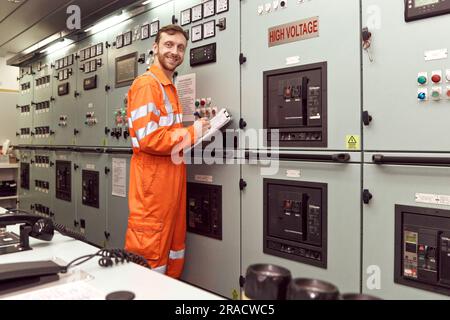 Jeune officier du génie inspectant le tableau électrique dans la salle de contrôle des moteurs. Ingénieur offshore souriant. Banque D'Images