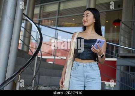 Jeune femme asiatique à la mode marchant dans un centre commercial avec un smartphone à la main. Une femme vietnamienne joyeuse a fait du shopping avec un sac fourre-tout Banque D'Images