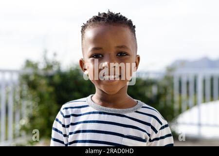 Portrait de mignon garçon afro-américain souriant à la caméra tout en se tenant contre le ciel clair dans la cour Banque D'Images