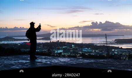 Homme qui prend des photos à l'aube à l'aide d'un smartphone au sommet du Mont Eden. Rangitoto Island au loin. Auckland. Banque D'Images