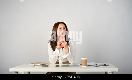 Une jeune femme de bureau asiatique belle et rêveuse tient son smartphone, regardant l'espace vide avec des yeux rêvants, et rêvant abo Banque D'Images