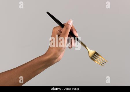 Main d'une femelle noire anonyme tenant une fourchette dorée avec une poignée noire sur fond gris Banque D'Images