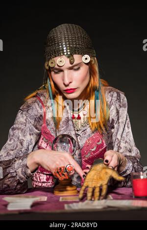 Une femme au grand renfeur de fortune en costume tsigane et coiffures montrant la prédiction de l'avenir sur la paume artificielle sur fond sombre Banque D'Images