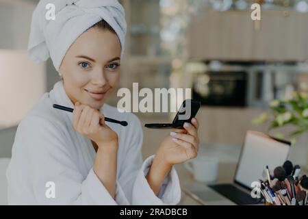 Une jeune femme applique du maquillage, pose dans un peignoir, avec un pinceau, dans un appartement moderne. Banque D'Images