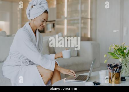 Indépendant travaille à la maison dans un peignoir, types sur ordinateur portable, boissons café, aime l'atmosphère domestique, surfer sur le Web, lit les conseils de soin de la peau. Banque D'Images