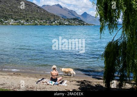 Une femme en train de bronzer avec un chien sur la plage de Frankton surplombant le lac Wakatipu près de Queenstown dans l'île du Sud de la Nouvelle-Zélande. Photo : Rob Watkins Banque D'Images