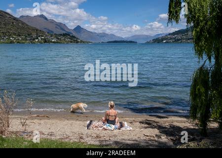 Une femme en train de bronzer avec un chien sur la plage de Frankton surplombant le lac Wakatipu près de Queenstown dans l'île du Sud de la Nouvelle-Zélande. Photo : Rob Watkins Banque D'Images