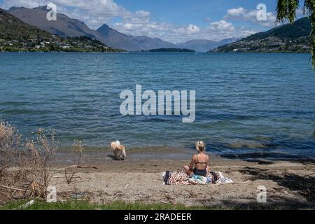 Une femme en train de bronzer avec un chien sur la plage de Frankton surplombant le lac Wakatipu près de Queenstown dans l'île du Sud de la Nouvelle-Zélande. Photo : Rob Watkins Banque D'Images