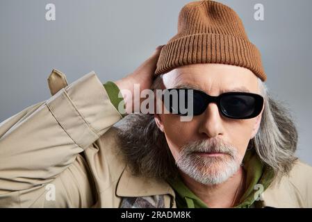 portrait de l'homme senior barbu et de style hipster en lunettes de soleil sombres, bonnet beanie et trench-coat beige tenant la main près de la tête et regardant l'appareil photo allumé Banque D'Images