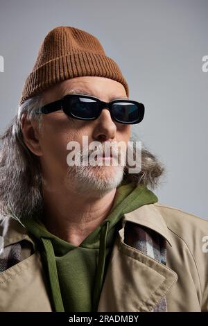 portrait de l'homme senior à poil gris et barbu en bonnet beanie, lunettes de soleil sombres et trench-coat beige regardant loin sur fond gris, taille basse Banque D'Images