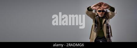 style hipster tendance homme en lunettes de soleil sombres et trench-coat beige posant avec les mains tendues sur fond gris, âge élégant, mode s. Banque D'Images