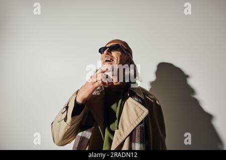 concept de vieillissement heureux et stylé, homme senior de style hippster excité en lunettes de soleil sombres, bonnet beanie et trench-coat beige regardant loin et riant sur gr Banque D'Images