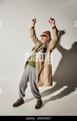 pleine longueur de style hippster expressif homme senior en lunettes de soleil sombres, bonnet beanie, trench-coat beige et écharpe à carreaux posant avec les mains levées sur le gris Banque D'Images