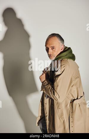 mode et âge, style personnel, homme senior confiant avec une expression sérieuse du visage debout dans un trench-coat beige et regardant la caméra sur fond gris Banque D'Images