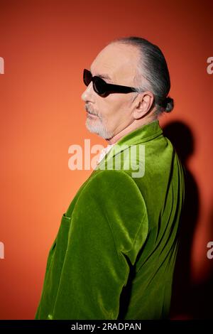 vue latérale de l'homme âgé à poil gris et à barbe en lunettes de soleil sombres et blazer en velours vert debout et regardant loin sur fond rouge et orange avec Banque D'Images