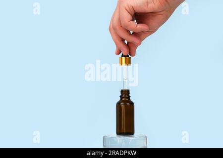 La main mâle maintient le compte-gouttes avec de l'huile. Ouvert pot de produits cosmétiques naturels sur podium en verre sur fond bleu. Huile essentielle pour le soin de la peau. Soins de la peau, Banque D'Images