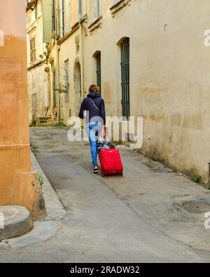 Jeune femme, marchant, tirant une valise roulante rouge dans l'étroite rue de la vieille ville sur Arles, France, Banque D'Images