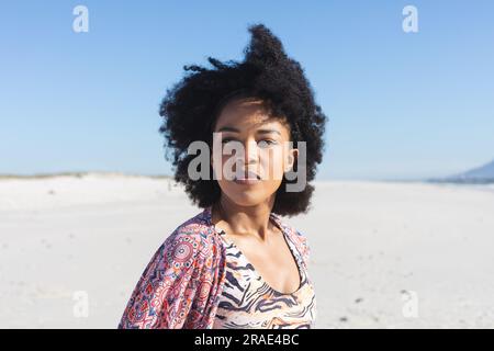 Femme afro-américaine réfléchie assise sur la plage ensoleillée regardant loin Banque D'Images