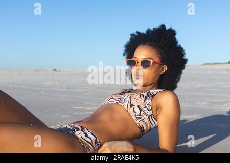 Heureuse femme afro-américaine en lunettes de soleil et bikini couchée sur la plage ensoleillée Banque D'Images