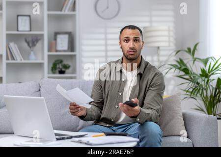 Portrait d'un jeune homme afro-américain inquiet assis sur le canapé à la maison, tenant un téléphone et des documents. Comptes chèques, a des problèmes financiers et de la dette. Il regarde malheureusement la caméra. Banque D'Images