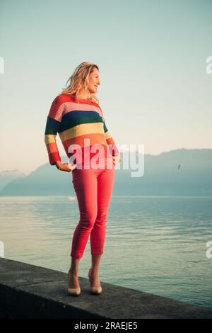 Belle femme se reposant au bord du lac au coucher du soleil le soir chaud, portant un pull-over coloré Banque D'Images