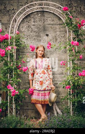 Portrait d'été de jolie petite fille portant une robe colorée, tenant un chapeau, posant dans un petit jardin de roses Banque D'Images