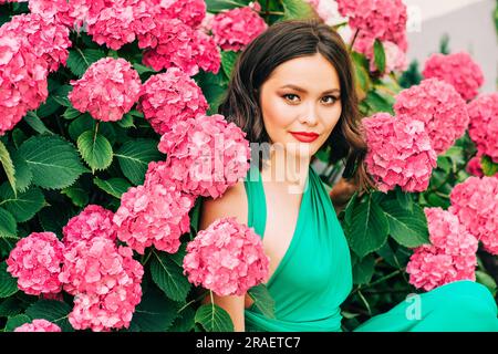 Portrait extérieur de la belle jeune femme posant dans des fleurs d'hortensia rose, portant une robe verte Banque D'Images