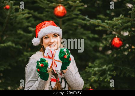 Portrait de fête de la jeune femme heureuse se posant à l'extérieur près de l'arbre de Noël, portant des moufles vertes et un chapeau de père Noël Banque D'Images