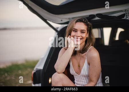 Jeune femme assise dans un coffre de voiture près du lac, profitant de l'été. Banque D'Images