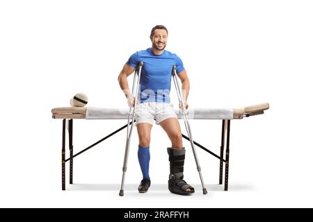 Joueur de football avec béquilles assis sur un lit de physiothérapie isolé sur fond blanc Banque D'Images