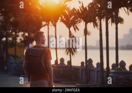 Vue arrière d'un jeune homme profitant de la vue sur la ville au beau coucher du soleil. Tourisme avec sac à dos à Hanoi, Vietnam. Banque D'Images