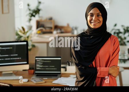 Jeune travailleur de bureau musulman souriant dans un hijab noir et un maillot rouge regardant la caméra tout en se tenant contre le lieu de travail avec des ordinateurs Banque D'Images