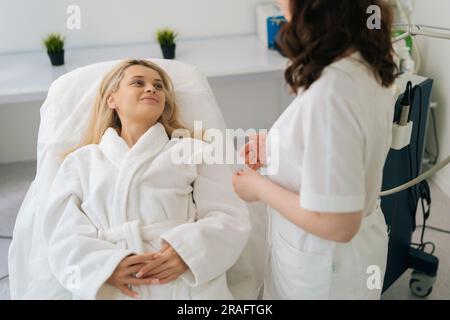 Vue en grand angle d'une femme blonde souriante et attrayante discutant avec une femme cosmetologiste avant la procédure de matériel de cosmétologie dans une clinique de beauté moderne. Banque D'Images