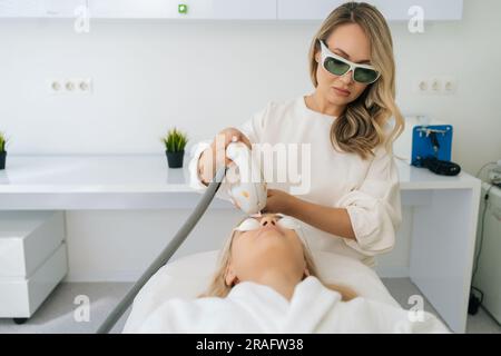 Portrait de cosmétologue dans des lunettes de protection exécutant la photothérapie anti-acné avec l'équipement. Belle femme dans le salon de beauté pendant la photo Banque D'Images