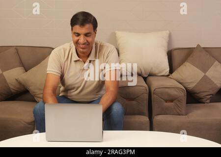 Homme indien heureux travaillant de la maison sur un ordinateur portable assis sur un canapé. Banque D'Images