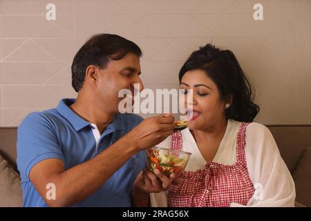 Heureuse femme de mari consciente de la santé indienne assise ensemble ayant un bol de salade verte assis sur le canapé dans un salon Banque D'Images