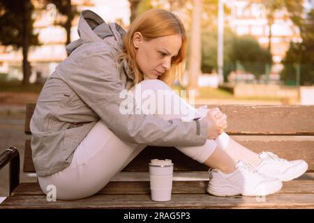 Une fille pensive d'apparence européenne aux cheveux blonds est assise sur un banc, une fille est assise dans une veste grise et un pantalon de survêtement blanc et des baskets. ph de haute qualité Banque D'Images