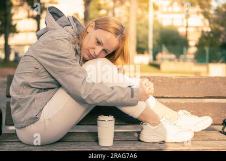 une fille aux cheveux blonds est assise sur un banc avec ses pieds et a abaissé sa tête à ses pieds, la fille est assise dans une veste grise et un pantalon de survêtement blanc, il y a un Banque D'Images