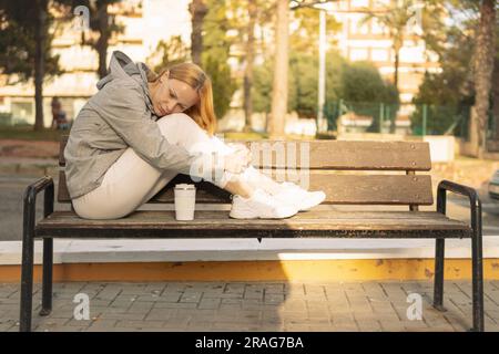 fille triste est assise sur un banc avec ses pieds et la tête abaissée à ses pieds, la fille est assise dans une veste grise et des pantalons de survêtement blancs et des baskets, là Banque D'Images