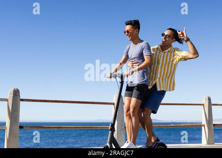 Heureux couple masculin gay biracial ayant du scooter d'équitation amusant ensemble sur la promenade par la mer, espace de copie Banque D'Images