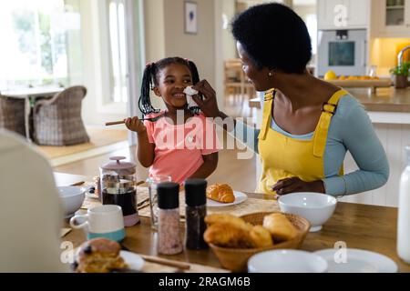 Mère afro-américaine nettoyant le visage de sa fille avec du papier de soie tout en prenant son petit déjeuner à la maison Banque D'Images