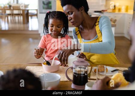 Mère afro-américaine versant du lait dans des céréales de petit déjeuner pour fille à la table à manger à la maison Banque D'Images