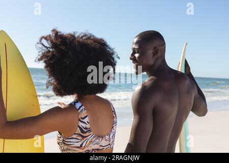 Heureux couple afro-américain avec des planches de surf debout sur la plage ensoleillée parlant Banque D'Images