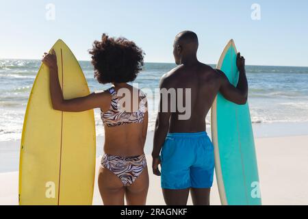 Vue arrière du couple afro-américain en forme tenant des planches de surf debout sur la plage ensoleillée au bord de la mer Banque D'Images