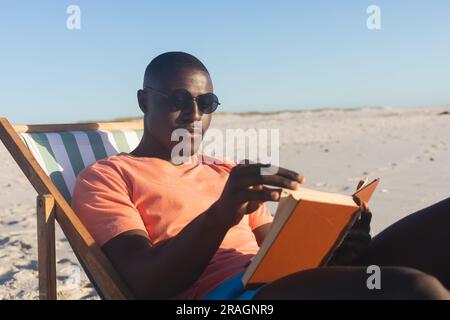 Heureux homme afro-américain dans des lunettes de soleil assis dans le transat lisant le livre sur la plage ensoleillée Banque D'Images