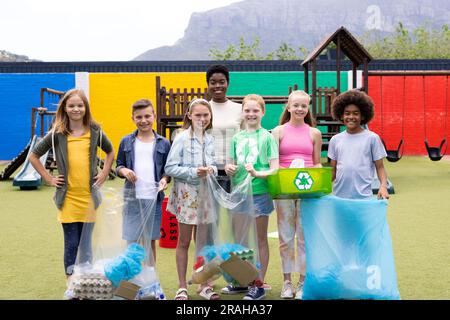 Portrait d'une enseignante et d'un élève joyeux et divers tenant recyclage collectés dans la cour d'école. Éducation, inclusion, école primaire et apprentissage Banque D'Images