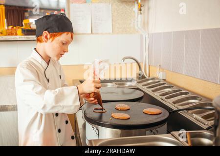 garçon cuisine des crêpes sur une crêperie professionnelle industrielle, cuisine dans le café, petit chef. Verse la pâte sur le poêle. Banque D'Images