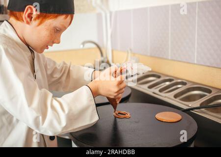 garçon cuisine des crêpes sur une crêperie professionnelle industrielle, cuisine dans le café, petit chef. Déverse la pâte de l'enveloppe de cuisson. Banque D'Images