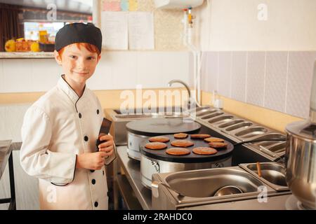 garçon cuisine des crêpes sur une crêperie professionnelle industrielle, cuisine dans le café, petit chef. enfant roux en costume de cuisinier. Banque D'Images